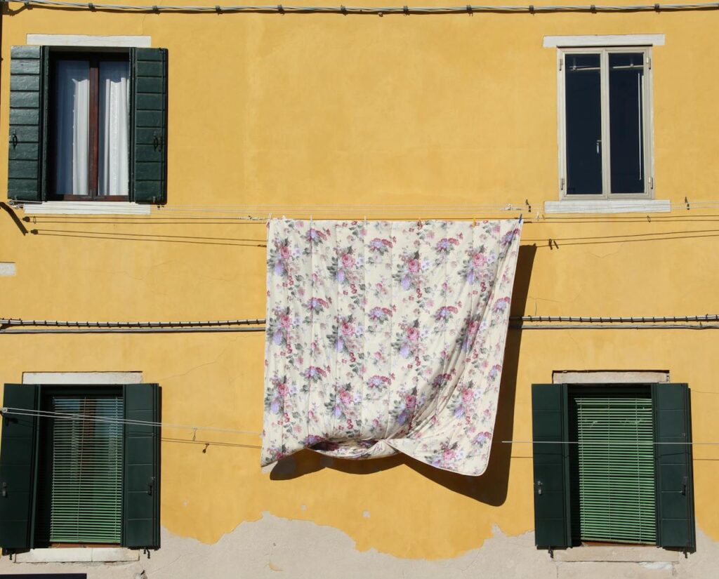 pexels photo 11148259 A floral blanket dries on a vibrant yellow building facade with green shutters and windows.